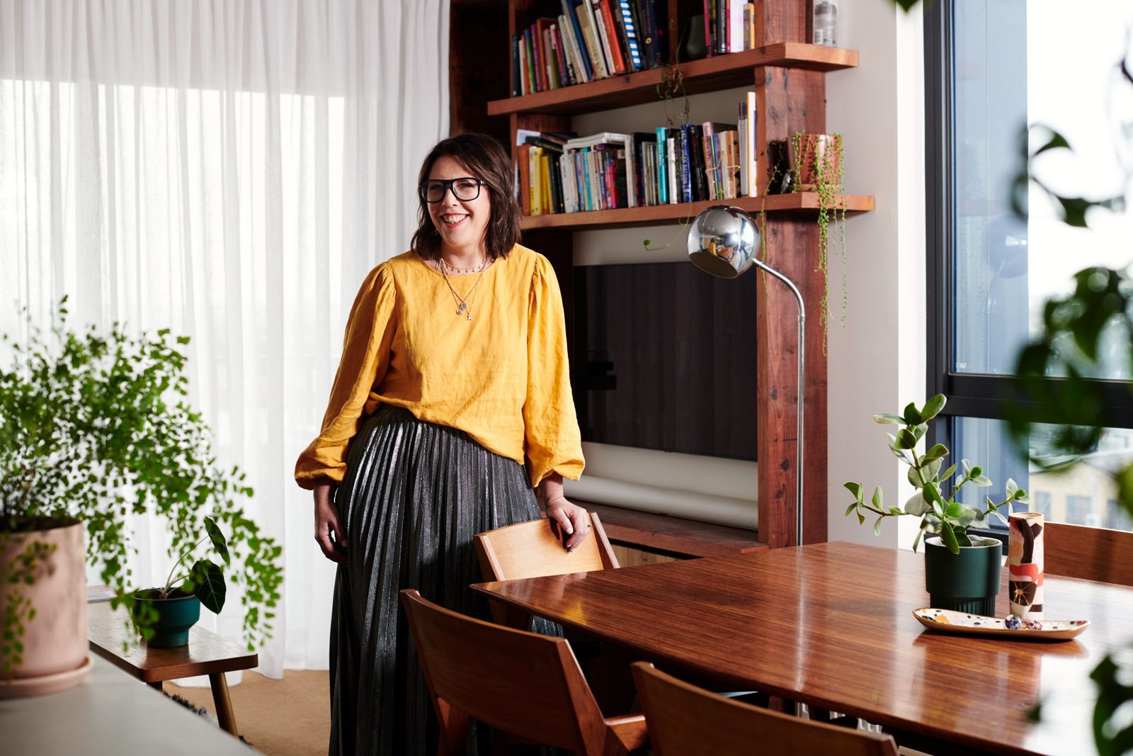 Karla in a yellow top in her apartment, beside a timber bookshelf and dining table