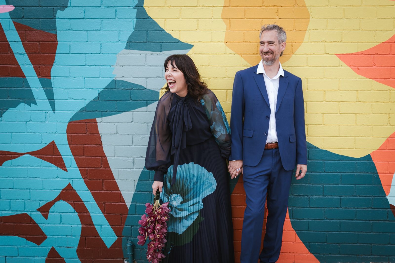 Karla laughing and Yanni smiling, holding hands in front of a colourful mural
