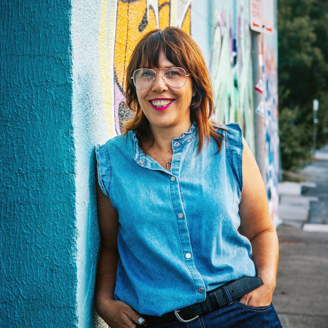 Karla leaning on a teal wall in Brunswick, smiling to camera