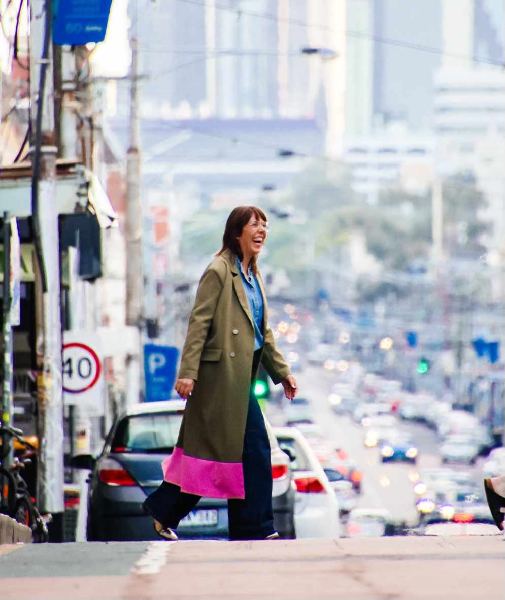 Karla laughing as she crosses a busy Brunswick street in a long olive coat
